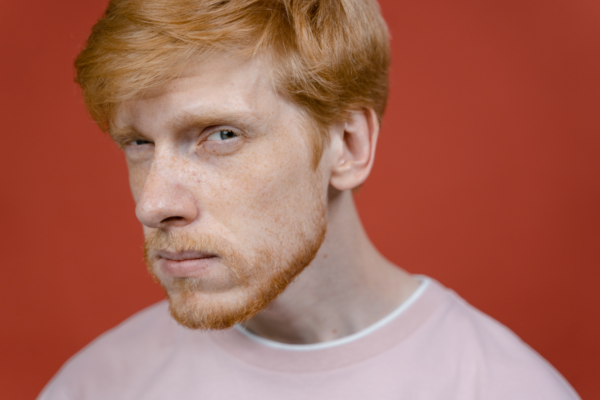 Homem branco, ruivo, dos olhos castanhos e usando uma camiseta rosa pastel, olhando para a câmera com expressão de dúvida, para demonstrar uma pessoa que não sabe o que é uma bio no Instagram. O fundo da imagem é marrom.