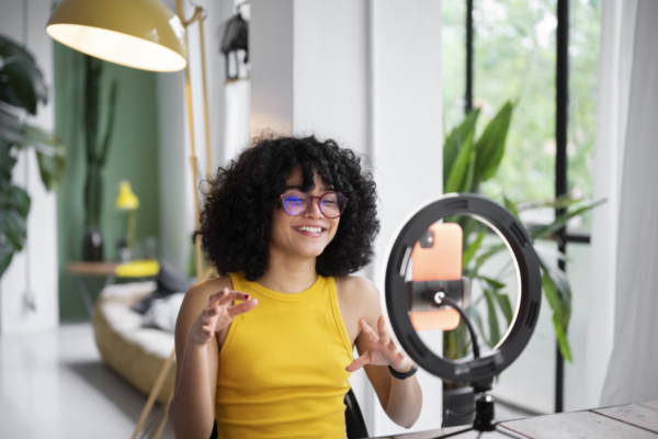 Mulher de cabelos cacheados volumosos, pretos e curtos, da pele clara, usando regata amarela. Ela trabalha com influencers, e está gravando conteúdo com o celular em uma ring light a sua frente.