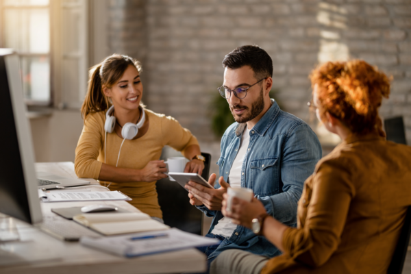 Na imagem, observamos três pessoas conversando em frente a uma mesa, com um monitor e vários cadernos dispostos acima. Eles aparentam estar discutindo estratégias de remarketing, tema do texto. Da esquerda para a direita, sentados, temos: uma mulher branca, do cabelo longo amarrado em um rabo de cavalo, vestindo um suéter bege e com um headset branco pendurado no pescoço; um homem branco de cabelo curtinho, vestindo uma camiseta branca e uma jaqueta jeans clara, segurando um tablet nas mãos; e, por último, uma mulher branca de costas, com o cabelo ruivo preso em um coque, vestindo uma blusa de manga longa marrom e segurando uma xícara de café.