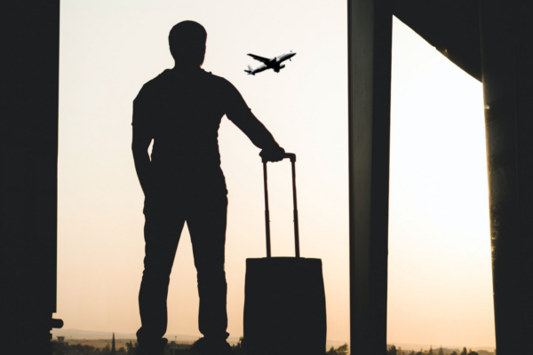 A foto mostra a silhueta de um homem em pé, segurando uma mala de rodinhas ao seu lado, observando o pôr-do-sol com um avião passando no céu.