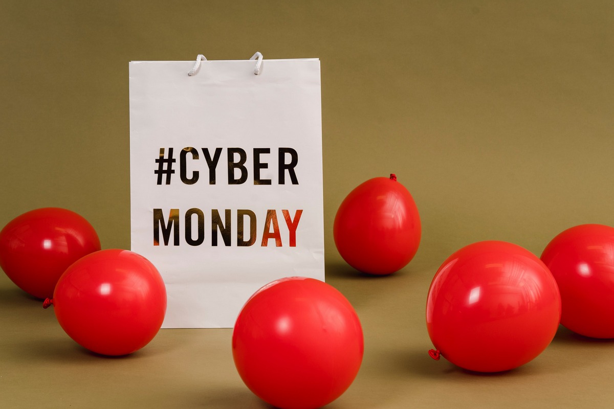 A imagem mostra uma sacola escrito #CYBERMONDAY, com várias bexigas vermelhas espalhadas pelo ambiente.