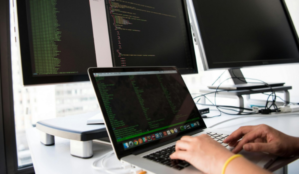 Aqui temos três telas ligadas de monitores, e uma pessoa teclando em um notebook disposto sobre a mesa. Subentende-se que ela está programando.