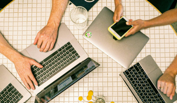 A imagem tem a vista de cima de uma mesa, com três notebooks abertos e um fechado. Entende-se que há quatro pessoas reunidas, todas apoiadas em seus aparelhos, estudando sobre transformação digital. Uma delas, apoiada no notebook fechado, segura um celular em suas mãos. Também encontramos um copo de vidro com água e um caderno com a capa preta dispostos sobre a mesa.