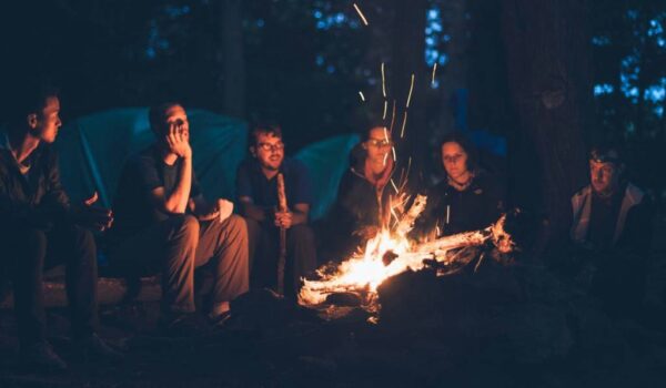 A imagem mostra um grupo de adultos em volta de uma fogueira, fazendo referência ao Turismo de Base Comunitária.