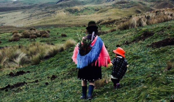 A imagem mostra duas pessoas no campo, uma adulta e uma criança, usando roupas típicas e carregando ramos de flores nas costas.