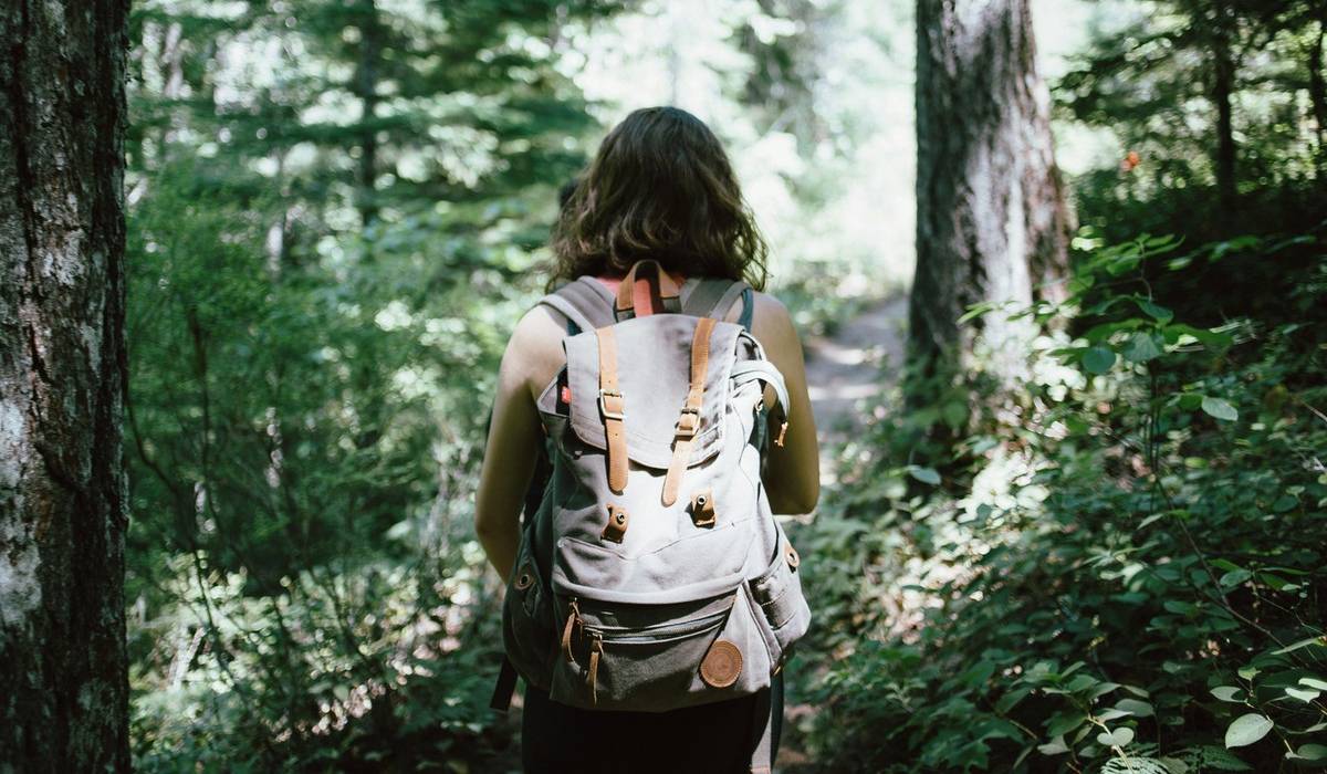 A imagem mostra uma mulher de costas andando em uma mata. Ela tem cabelo longo e está de mochila.